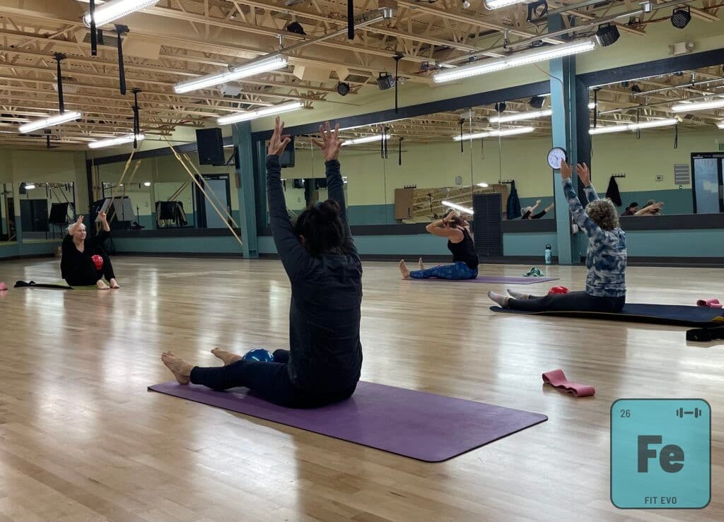 A member performing a seated roll-back with arms overhead during a Pilates class.