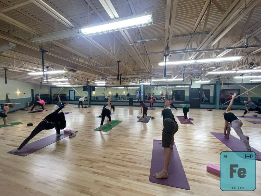 A yoga class in the FIT EVO studio, members spaced out on mats across the wood floor holding a standing triangle pose, with the mirrored wall behind them.