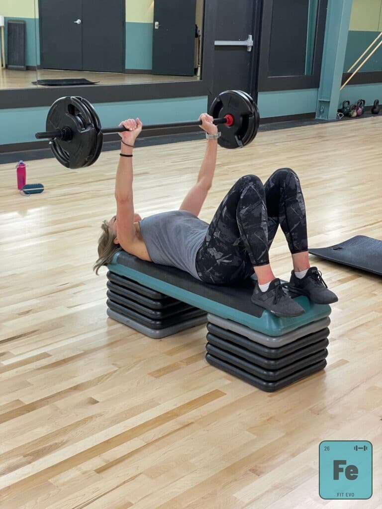 A member lying on a step bench pressing a barbell overhead in the FIT EVO studio.