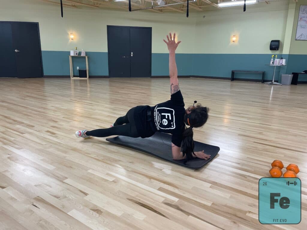 An instructor on a mat performing a lying rotation with one arm reaching upward.