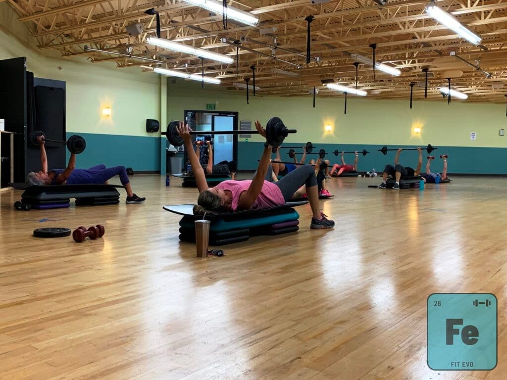 Members lying on step benches pressing barbells during a Group Power class.