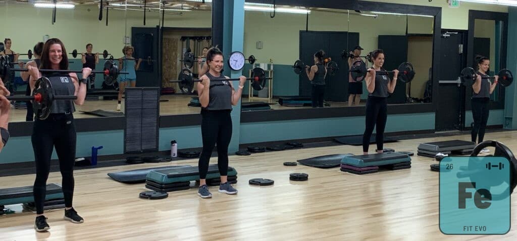 Members standing with barbells racked at the shoulder during a class.