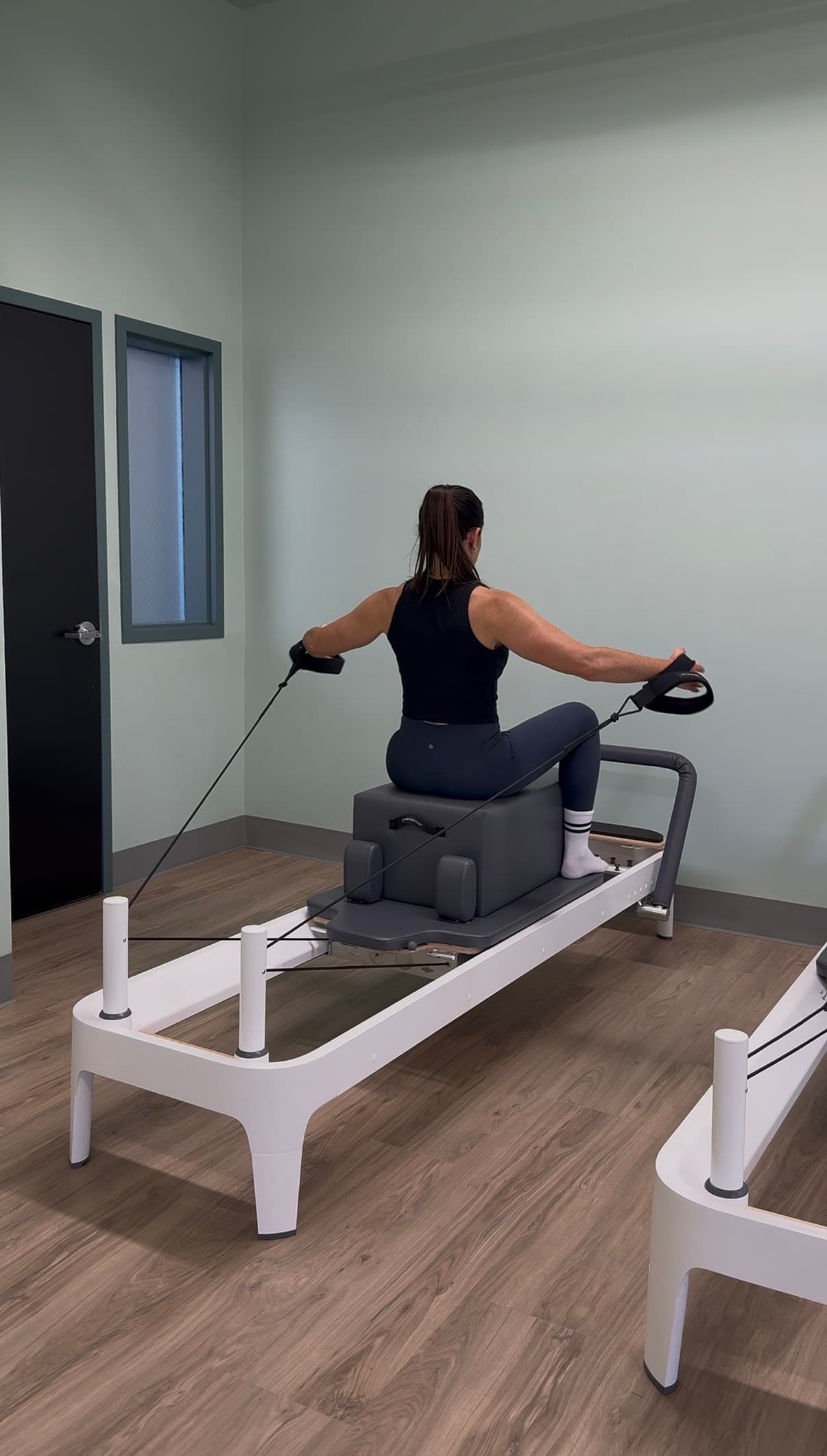 A Fit Evo instructor seated on the reformer box with both arms extended against the straps, in the Bellingham reformer studio